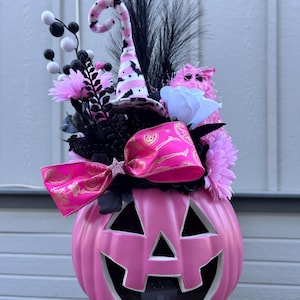 May include: A pink Halloween pumpkin with a carved face, filled with black and pink decorative elements. The arrangement includes a witch hat, flowers, feathers, and a pink bow with gold accents. A festive centerpiece for the holiday.