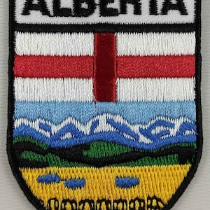 May include: Embroidered patch featuring the Alberta provincial flag. The flag is a blue, white, and green shield with a red cross and a yellow field at the bottom. The word "ALBERTA" is embroidered in black on a white background at the top of the patch.