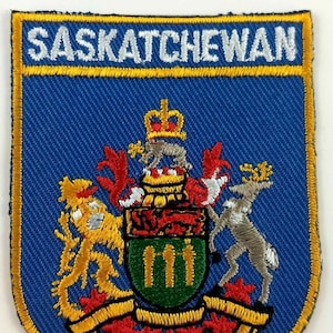 May include: A blue and gold embroidered patch with the word "SASKATCHEWAN" at the top and the coat of arms of Saskatchewan in the center. The coat of arms features a lion, a unicorn, and a shield with three green stripes.