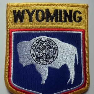 Peut inclure: Un écusson brodé jaune avec le mot "WYOMING" en lettres noires. L'écusson présente un fond bleu avec une silhouette blanche de bison et un emblème circulaire au centre.