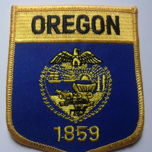 May include: A blue and gold embroidered patch featuring the state seal of Oregon. The seal depicts a golden eagle, a wagon, and the year 1859. The words "OREGON" are embroidered in black on a gold background above the seal.