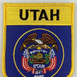 May include: A blue and yellow embroidered patch with the state seal of Utah. The seal features a bald eagle, a beehive, and the words "Industry" and "Utah 1847 1896". The patch has a yellow border and the word "Utah" in black letters at the top.