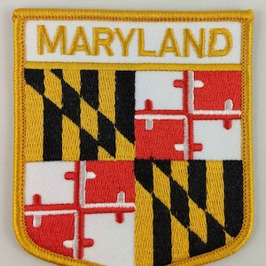 Puede incluir: Un parche bordado amarillo con la bandera del estado de Maryland. La bandera presenta un escudo cuartelado en negro y oro con cuatro cruces rojas sobre fondo blanco. La palabra "MARYLAND" está bordada en amarillo en la parte superior del parche.