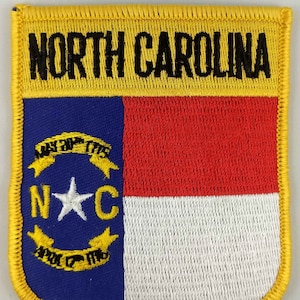 Könnte beinhalten: Ein gelber gestickter Aufnäher mit den Worten "NORTH CAROLINA" in schwarzen Buchstaben. Der Aufnäher zeigt die Staatsflagge von North Carolina, die ein blaues Feld mit einem weißen Stern und den Buchstaben "NC" in Weiß enthält, umgeben von zwei gelben Schriftrollen mit den Worten "MAY 20, 1775" und "ANNO DOMINI 1776". Die Flagge enthält auch einen roten und weißen Streifen.