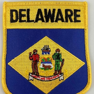 以下が含まれることがあります： 黄色と青色の刺繍パッチで、黒い文字で「DELAWARE」と書いてあり、中央にはデラウェア州の州章があります。