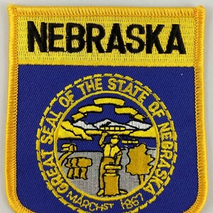 May include: A blue and gold embroidered patch featuring the state seal of Nebraska. The seal depicts a tree, a building, and a man holding a plough. The text "Great Seal of the State of Nebraska" and "March 1st 1867" are visible on the seal. The word "Nebraska" is embroidered in black on a yellow background at the top of the patch.