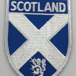 May include: Embroidered patch with the Scottish flag design. The patch has a blue background with a white St. Andrew's cross. The word "SCOTLAND" is embroidered in white at the top. A white lion rampant emblem is at the bottom.