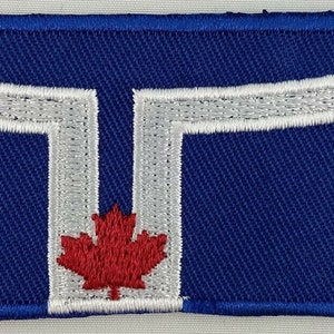 Può includere: Toppa ricamata con una forma a "T" bianca su sfondo blu. Una foglia d'acero rossa è centrata nella forma a "T".