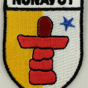 May include: An embroidered patch featuring the coat of arms of Nunavut, Canada. The patch is yellow, white, and red with a black outline. The coat of arms features an inukshuk, a traditional Inuit stone structure, and a blue five-pointed star.