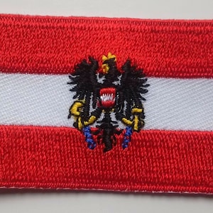 May include: A red, white, and black embroidered patch featuring the Austrian coat of arms, a black eagle with a golden crown and a red shield.