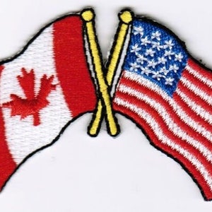 Peut inclure: Un drapeau canadien rouge et blanc et un drapeau américain rouge, blanc et bleu sont croisés sur des poteaux jaunes.
