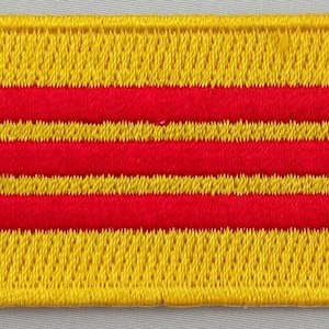 May include: A rectangular embroidered patch featuring a horizontal pattern of three red stripes alternating with four yellow stripes. The edges of the patch are finished with a thick yellow stitched border, creating a textured and durable appearance.