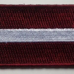 May include: A red and white embroidered patch featuring the flag of Latvia. The patch is made of a textured fabric and has a white border.