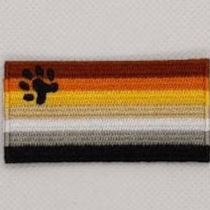 Può includere: Una bandiera arcobaleno di colore marrone, arancione, giallo, bianco, grigio e nero con un'impronta di zampa marrone nell'angolo in alto a sinistra.