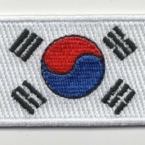 May include: A white embroidered patch with the South Korean flag. The flag features a white circle with a red and blue yin-yang symbol in the center, surrounded by four black trigrams.