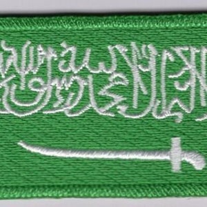 Puede incluir: Un parche bordado verde que representa la bandera de Arabia Saudita. El diseño incluye escritura árabe blanca y una espada blanca sobre un fondo verde. El parche tiene un borde cosido.