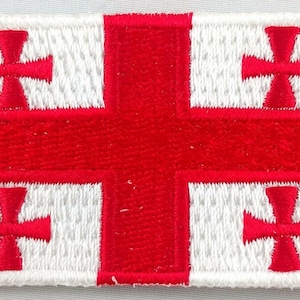 May include: A white embroidered patch with a red cross in the center and four smaller red crosses in the corners. The patch is a representation of the Georgian flag.