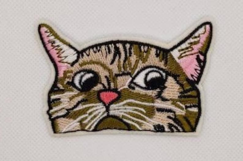 May include: Embroidered iron-on patch of a brown tabby cat with large, wide eyes and a surprised expression. The cat's nose and mouth are pink.