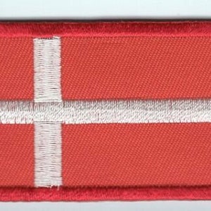 May include: A rectangular embroidered patch featuring a red background with a white Nordic cross design. The patch has a thick, stitched red border that frames the entire rectangular shape, creating a clean and defined edge for the fabric emblem.