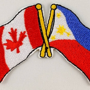 May include: Two embroidered flags, one Canadian and one Philippine, are crossed over each other. The Canadian flag is red and white with a red maple leaf. The Philippine flag is red, white, and blue with a yellow sun and three stars.