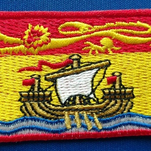 Könnte beinhalten: Gestickter Aufnäher mit der Flagge von New Brunswick, Kanada. Die Flagge ist rot und gelb mit einem goldenen Löwen und einem weißen Schiff auf einer blau-weißen Wellenlinie.