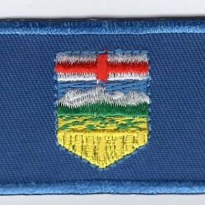 Può includere: Una toppa ricamata blu con lo stemma dell'Alberta, Canada. Lo stemma presenta uno scudo con una croce rossa su sfondo bianco, un cielo blu e bianco, montagne verdi, un campo giallo e un campo di grano giallo e bianco.