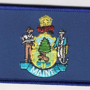 May include: A rectangular embroidered patch featuring the Maine state seal on a dark blue background. The design includes a central shield with a pine tree and a moose, flanked by a farmer and a sailor, topped with a star and the word MAINE.