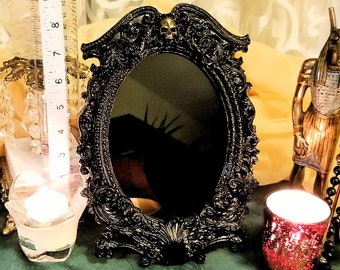 A skull is nestled atop this scrying mirror of gloss black or heavily antiqued silver with elegant leafy scrollwork in a heavy/thick frame