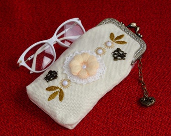 May include: A white eyeglass case with a floral design and a metal clasp. The case has a chain attached to it with a heart-shaped charm. The case is sitting on a red surface.