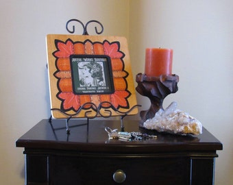 May include: A wooden picture frame with an orange, red, and brown design. The frame is on a black stand and has a picture of a woman with the text "Artful Works Boutique" inside. The frame is on a dark brown wooden table with a candle and a crystal cluster.