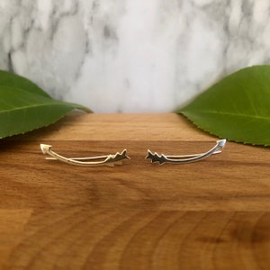 May include: A pair of silver arrow ear cuffs. The cuffs are curved and have a pointed arrowhead design.