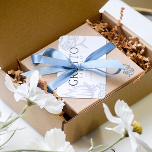 May include: A brown cardboard box with a blue ribbon tied around it. The box is filled with brown packing paper and a white card with the text "Grazie Catholic" on it.