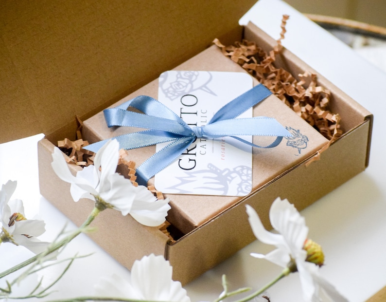 May include: A brown cardboard box with a blue ribbon tied around it. The box is filled with brown packing paper and a white card with the text "Grazie Catholic" on it.
