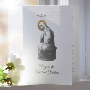 May include: A white card with a black and white illustration of the Virgin Mary. The card has a gold dove and halo. The text on the card reads "Prayers for Expectant Mothers".