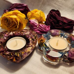 Op de afbeelding: Twee theelicht-kaarshouders gemaakt van helder hars met ingebedde gedroogde bloemen. De houders zijn gevormd als een hart en een bloem. De kaarsen zijn wit en branden.