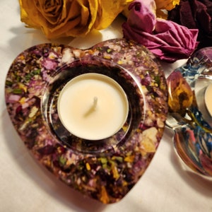 Op de afbeelding: Een hartvormige kaarsenhouder gemaakt van helder hars met ingebedde gedroogde bloemen in tinten van paars, roze en geel. Een witte theelichtkaars is in het midden van de houder geplaatst.