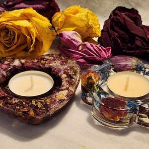Op de afbeelding: Twee hartvormige theelichthouders gemaakt van helder hars met ingebedde gedroogde bloemen en bloemblaadjes. De houders zijn gevuld met witte theelichtjes.