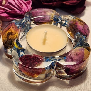 Op de afbeelding: Een glazen theelichthouder met een bloemmotief. De houder is gevormd als een bloem met meerdere bloemblaadjes. De bloemblaadjes zijn gevuld met gedroogde bloemen in tinten van paars, roze, geel en blauw. Een witte theelichtkaars brandt in het midden van de houder.