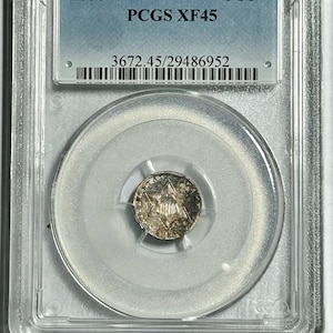 Puede incluir: Una moneda de plata de tres centavos de 1856, encapsulada en una funda protectora transparente. La moneda presenta un diseño de estrella y está calificada como PCGS XF45. La funda tiene una etiqueta azul con el año y la calificación.