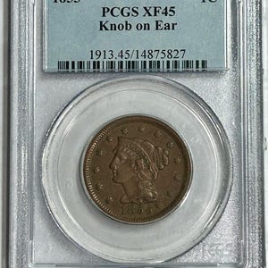 May include: A copper 1855 US large cent coin, graded PCGS XF45, with a "Knob on Ear" variety, encased in a clear protective holder. The coin features a profile of a woman and the text "1855".