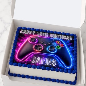 Könnte beinhalten: Ein rechteckiger Kuchen in einer weißen Schachtel mit einem essbaren Aufdruck eines schwarzen Game-Controllers mit neonpinken und blauen Umrissen vor einem Galaxie-Hintergrund. Der Text lautet HAPPY 10TH BIRTHDAY und JAMES, mit blauem Zuckerguss.