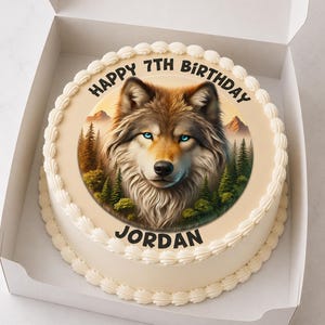 Könnte beinhalten: Ein runder Geburtstagskuchen in einer weißen Box. Der Kuchen zeigt ein detailliertes Bild eines Wolfes mit blauen Augen, umgeben von Bäumen und Bergen. Der Text "HAPPY 7TH BIRTHAY JORDAN" ist am Rand des Kuchens geschrieben.