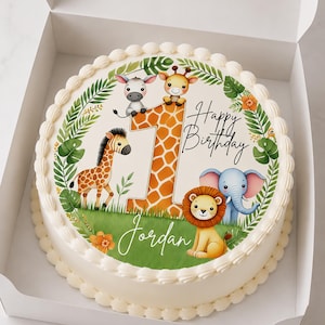 Könnte beinhalten: Ein runder Kuchen mit weißem Zuckerguss und einem dekorativen Rand. Der Kuchen ist mit Zeichentieren wie Zebra, Giraffe, Löwe und Elefant verziert. Die Zahl 1 ist abgebildet, zusammen mit den Worten "Happy Birthday" und einem Namen. Der Kuchen befindet sich in einer weißen Box.