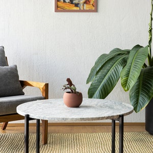 May include: A round marble coffee table with a black metal frame sits on a woven rug in a living room. A potted plant and a framed watercolor painting of cats eating ramen are visible in the background.