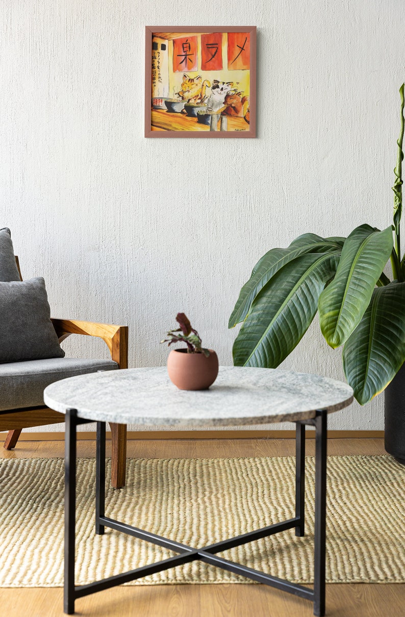 May include: A round marble coffee table with a black metal frame sits on a woven rug in a living room. A potted plant and a framed watercolor painting of cats eating ramen are visible in the background.