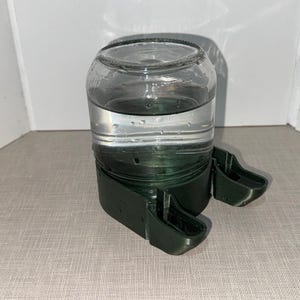 Könnte beinhalten: Ein klares Glasgefäß, gefüllt mit Wasser, steht auf einer dunkelgrünen Kunststoffbasis mit zwei trogartigen Ausbuchtungen. Das Glas ist teilweise mit Wasser gefüllt, und die Basis scheint das Gefäß sicher zu halten.
