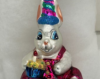 以下が含まれることがあります： ピンクとブルーの誕生日帽子をかぶった白いウサギが、火が灯ったろうそくがついた青いカップケーキを持っています。ウサギはピンクのオーバーオールを着ており、足元には緑の草と黄色の花があります。オーナメントはガラス製で、キラキラとした仕上げになっています。