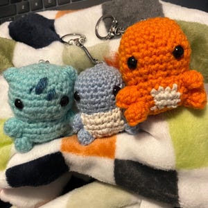 May include: Three crocheted plush toys with keychains. The first is a light blue, the second is a light blue with a white stripe, and the third is orange with a white stripe.