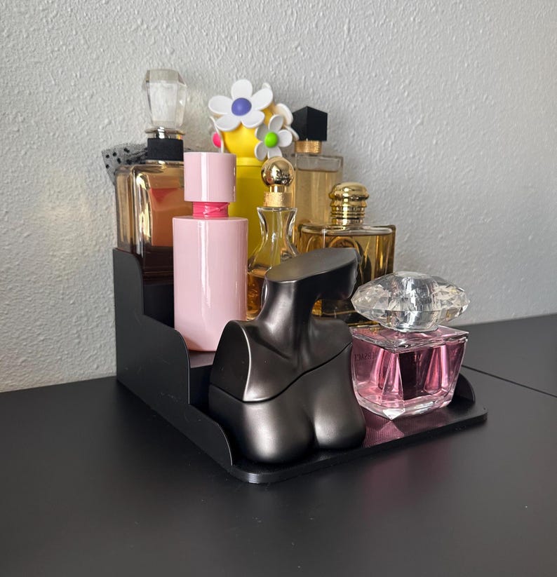 May include: A black tiered display stand with a variety of perfume bottles. The bottles are in various shapes and sizes, and some have decorative caps. The bottles are filled with different colored liquids, including pink, clear, and gold. The stand is on a black surface.
