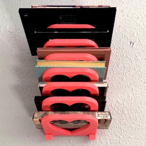 May include: A pink heart-shaped bookend holds a stack of books. The books are various sizes and colors, including black, brown, and blue. The bookend is made of plastic and has a smooth, glossy finish.
